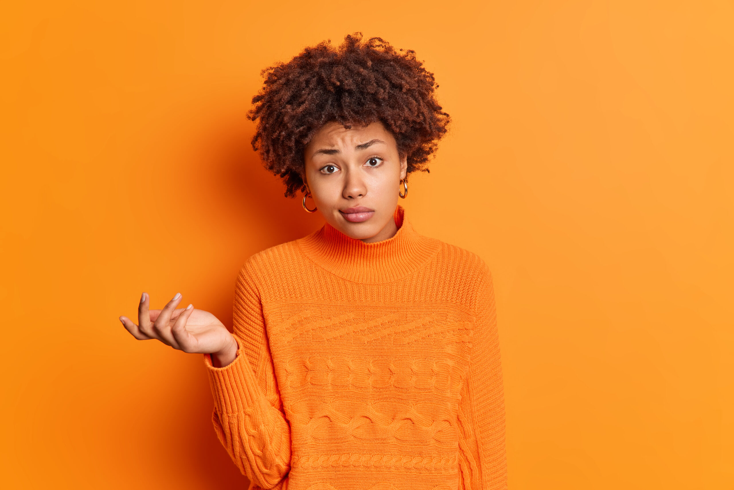 Horizontal shot of confused hesitant young Afro American woman shrugs shoulders looks perplexed and doubtful looks unconfident dressed in casual jumper isolated over vivid orange background. Horizontal shot of confused hesitant young Afro American woman shrugs shoulders looks perplexed and doubtful looks unconfident dressed in casual jumper isolated over vivid orange background.